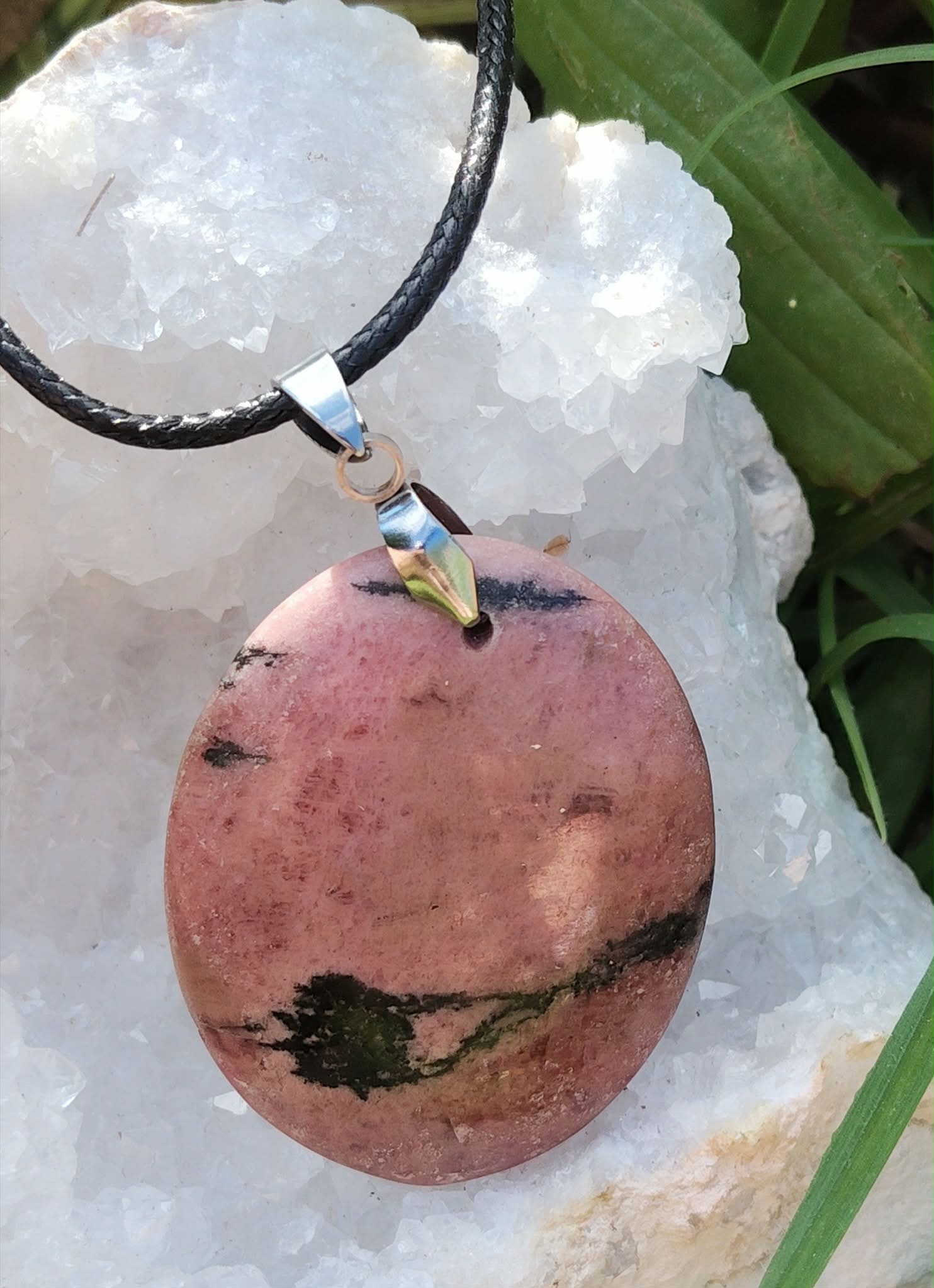Pendentif rond en rhodonite sur cordon noir, posé sur amas de quartz blanc et feuilles vertes, bijou en pierre naturelle relié à l’apaisement du cœur, à l’ancrage doux et à la sérénité.