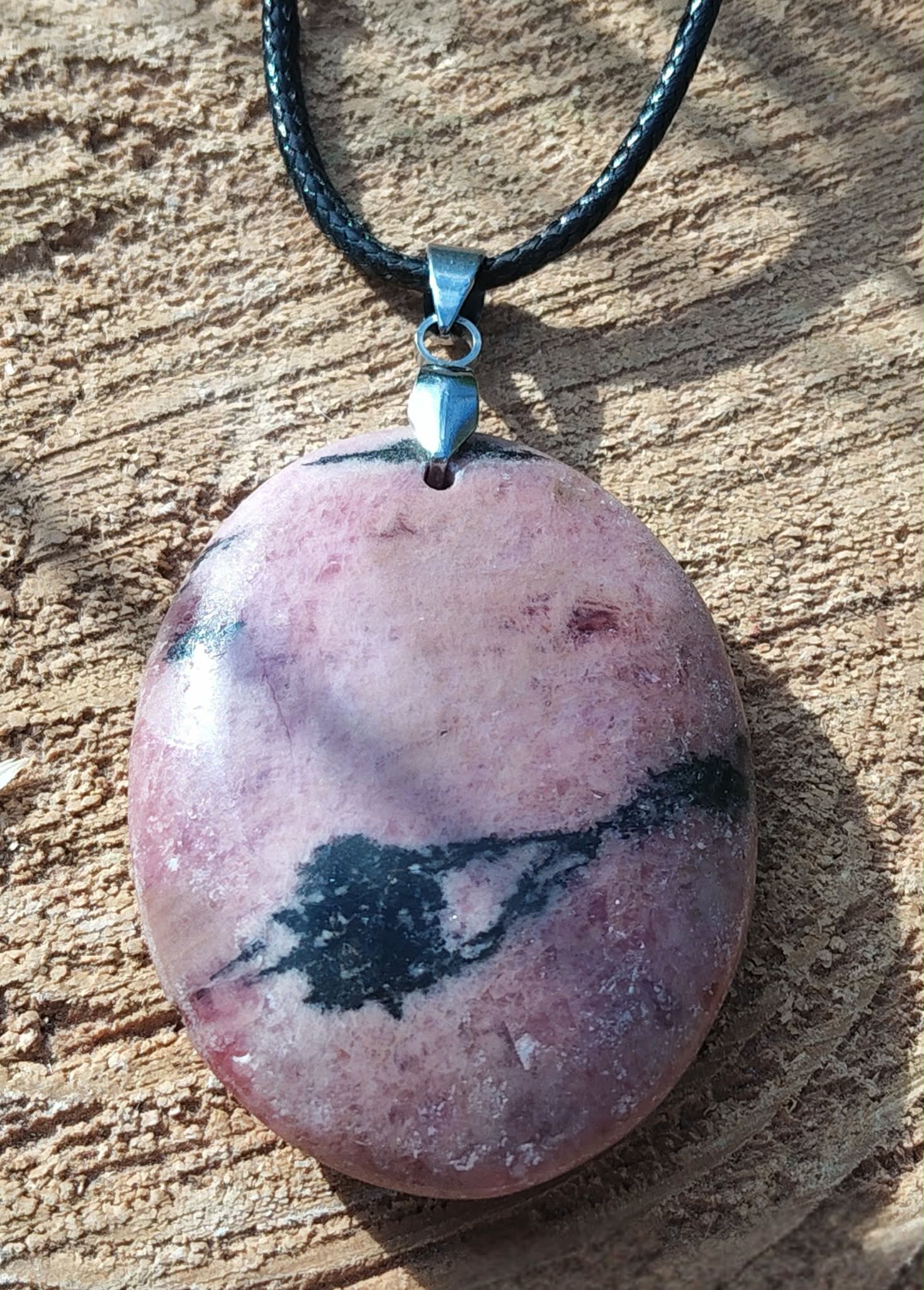 Pendentif ovale en rhodonite polie sur cordon noir, posé sur une tranche de bois au soleil, bijou en pierre naturelle évoquant le pardon, le recentrage et la paix émotionnelle.