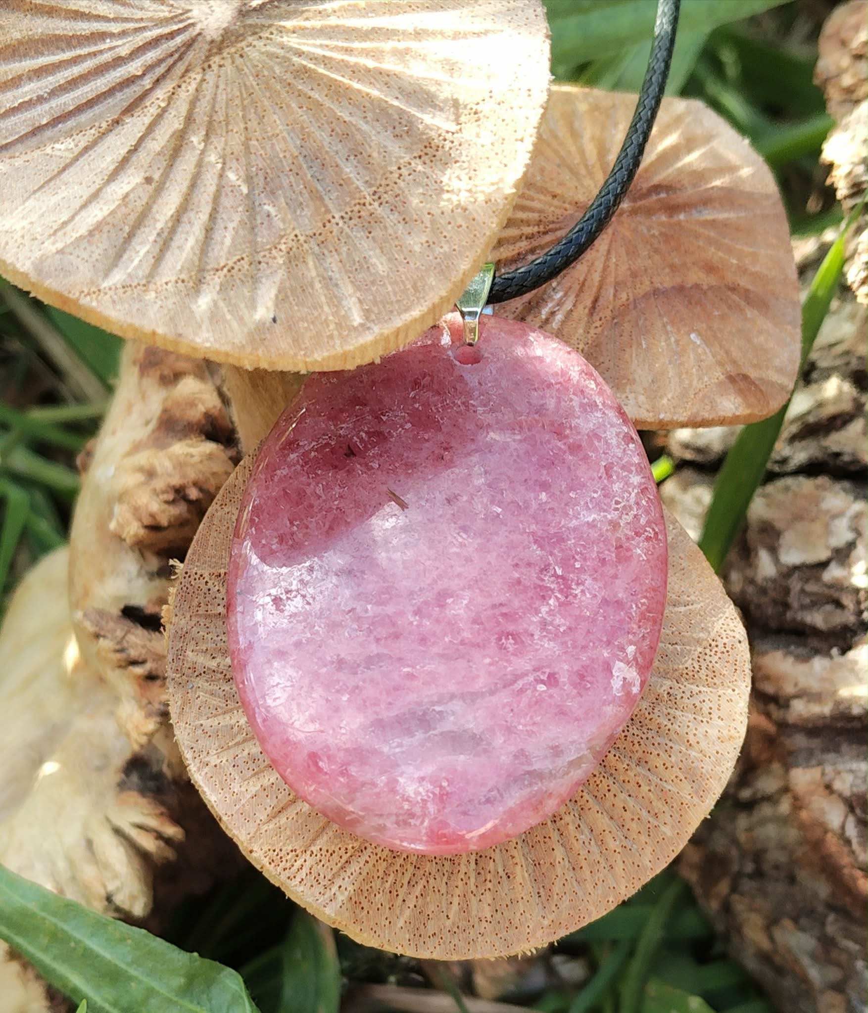 Pendentif rond en rhodonite sur cordon noir, posé sur champignons et herbes au soleil, bijou en pierre naturelle associé à la douceur, au réconfort affectif et à l’équilibre du cœur.