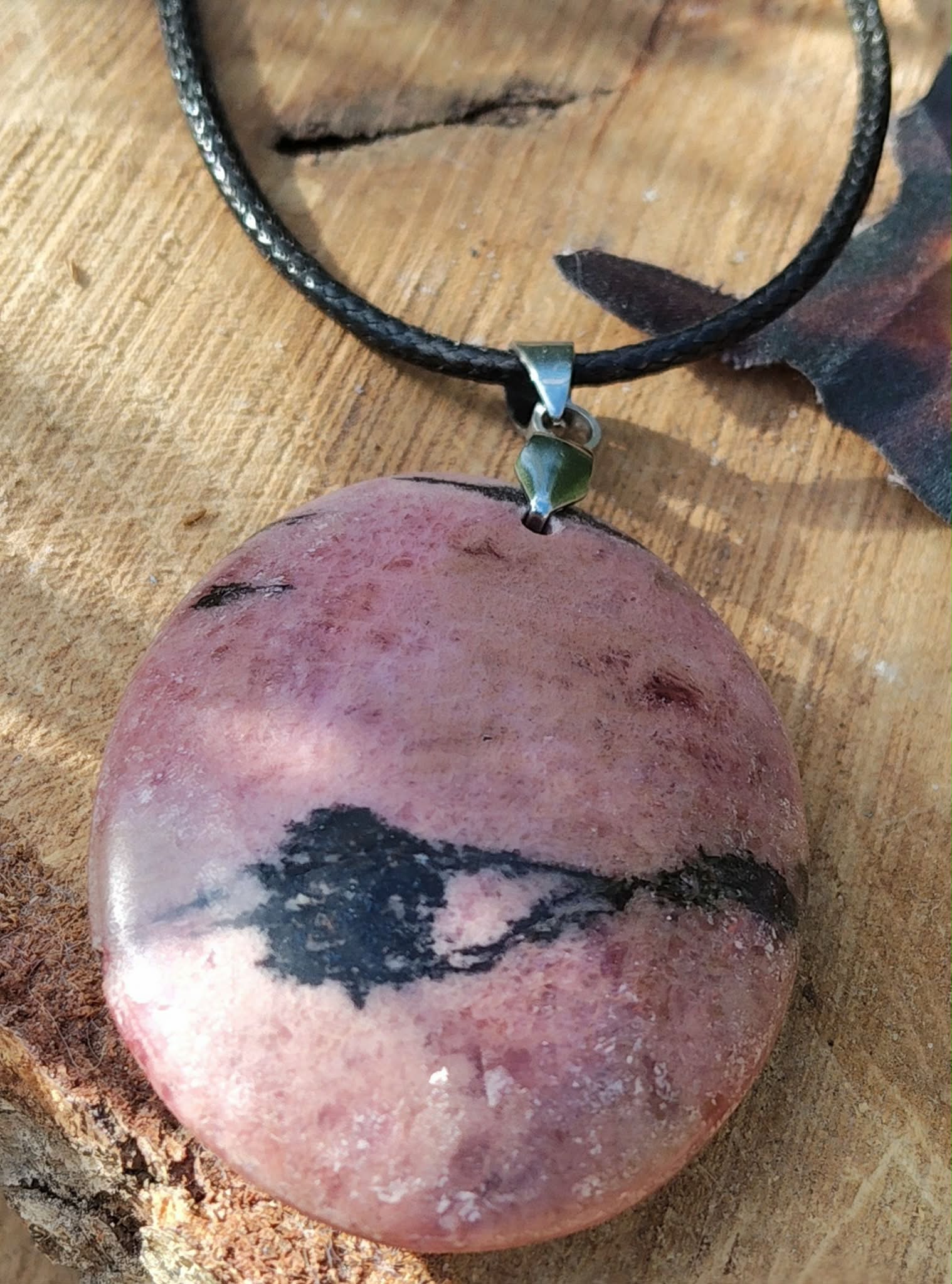 Pendentif rond en rhodonite rose sur cordon noir, posé sur une rondelle de bois, bijou en pierre naturelle évoquant le pardon, la douceur du cœur et la stabilité émotionnelle.