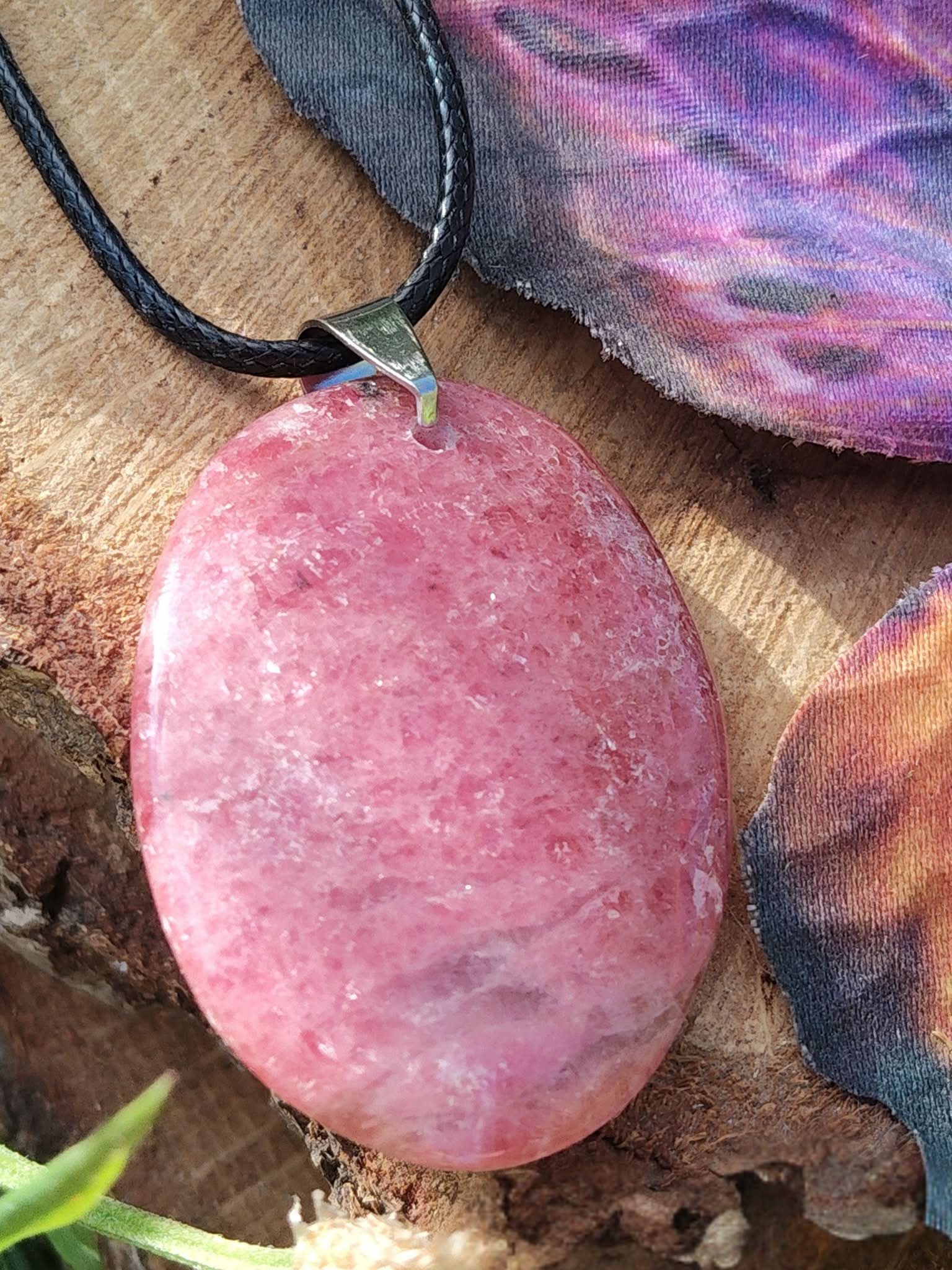 Pendentif ovale en rhodonite rose sur cordon noir, posé sur bois brut près d’une aile de papillon violette, bijou en pierre naturelle lié à la douceur intérieure, au soutien émotionnel et à la paix du cœur.