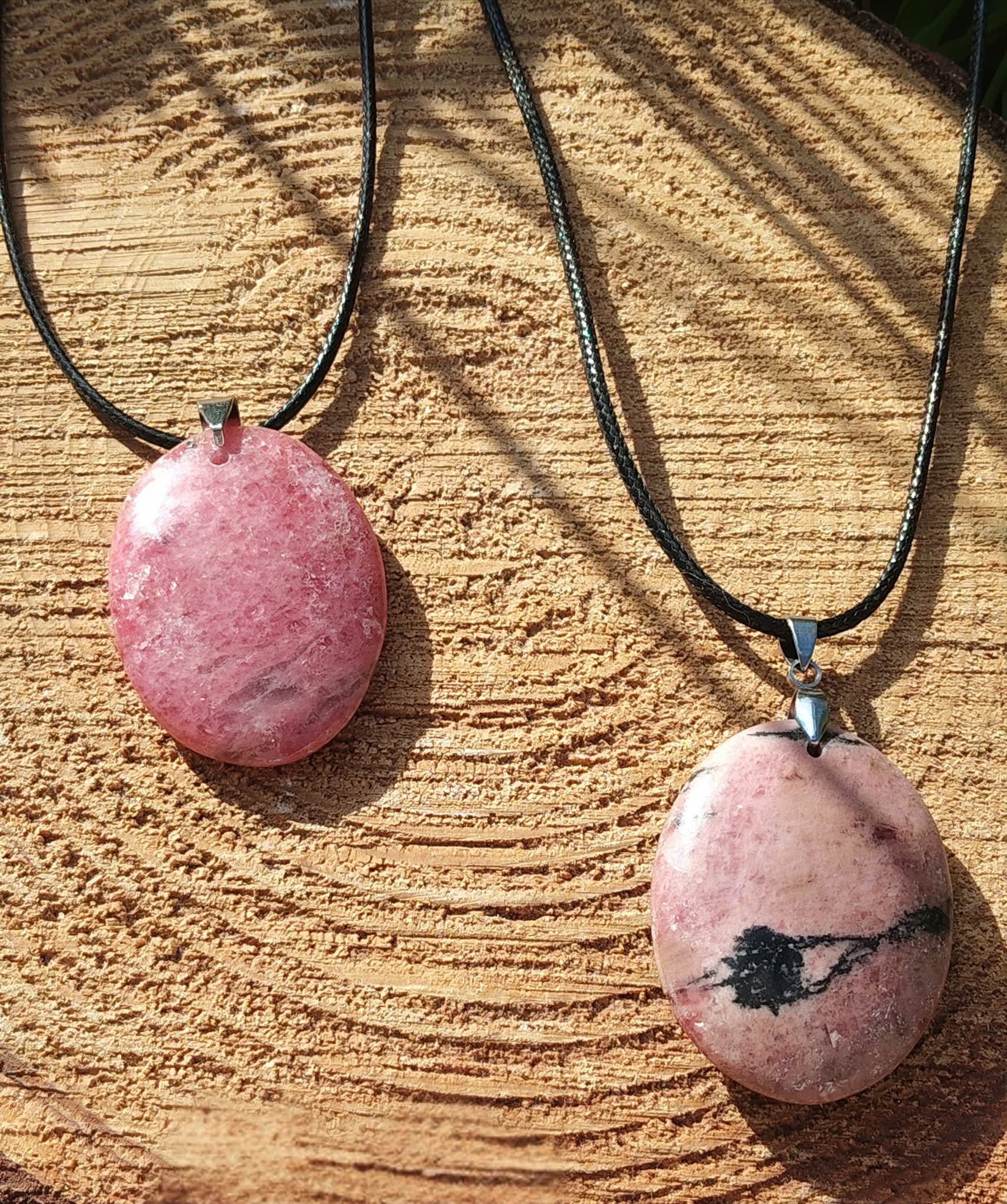Deux pendentifs en rhodonite sur cordon noir, posés sur une tranche de bois au soleil, bijoux en pierre naturelle associés à la douceur du cœur, au réconfort et à la stabilité émotionnelle.