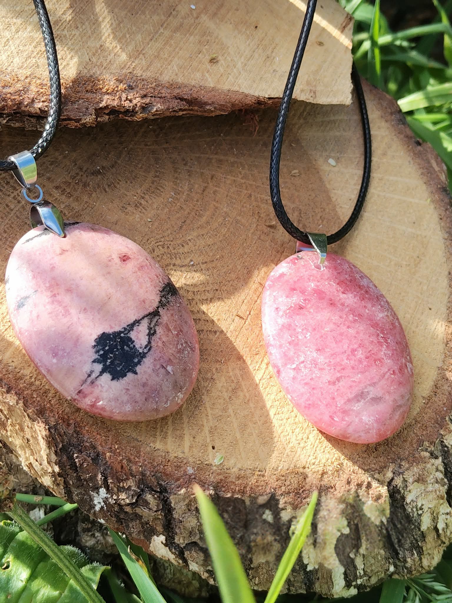 Deux pendentifs en rhodonite rose sur cordon noir, posés sur une rondelle de bois au jardin, bijoux en pierre naturelle associés à la tendresse, à l’équilibre affectif et au calme intérieur.