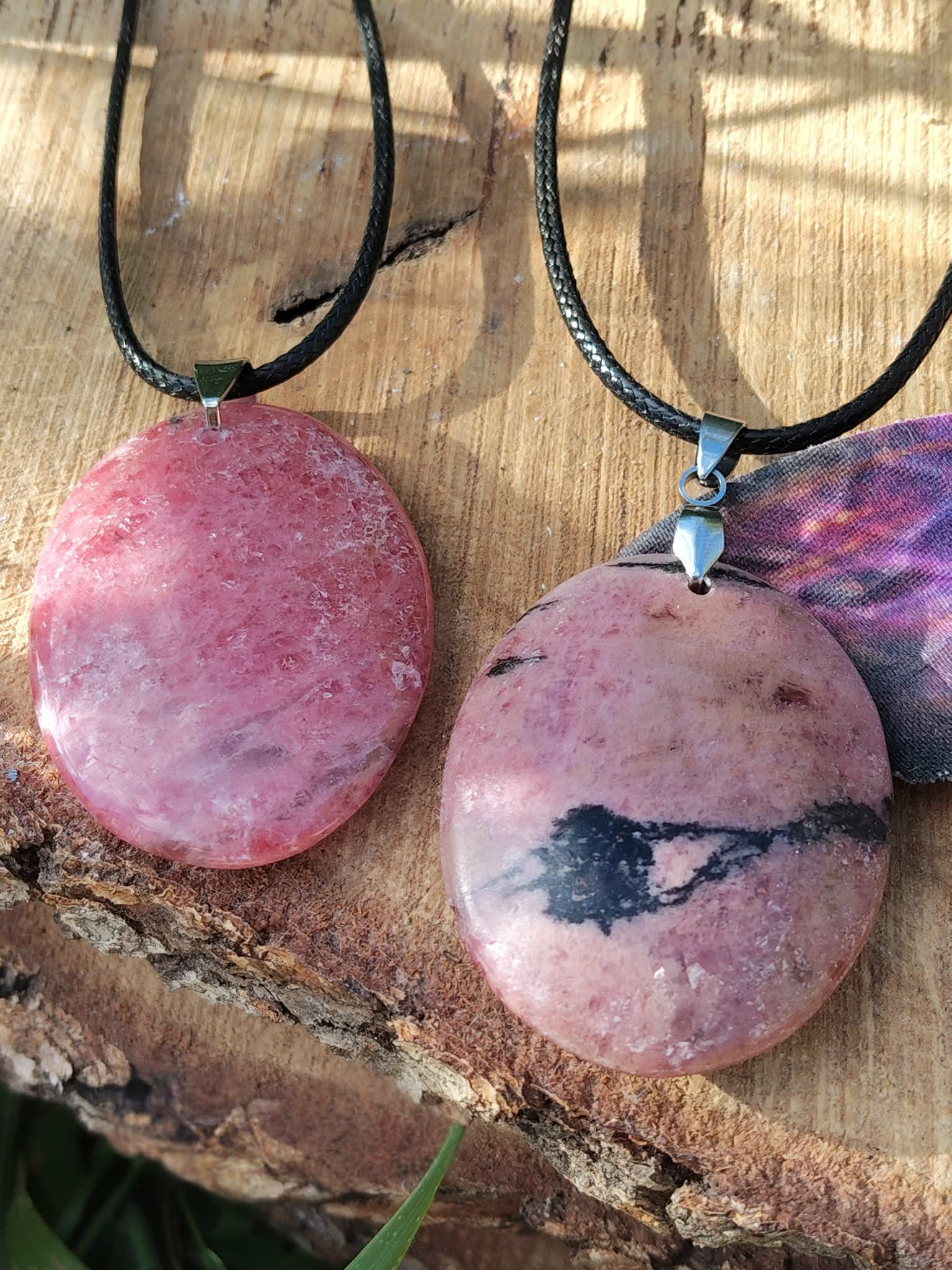 Deux pendentifs en rhodonite rose sur cordons noirs, posés sur écorce et plume violette, bijoux en pierre naturelle associés au réconfort, à l’harmonie émotionnelle et à la douceur relationnelle.