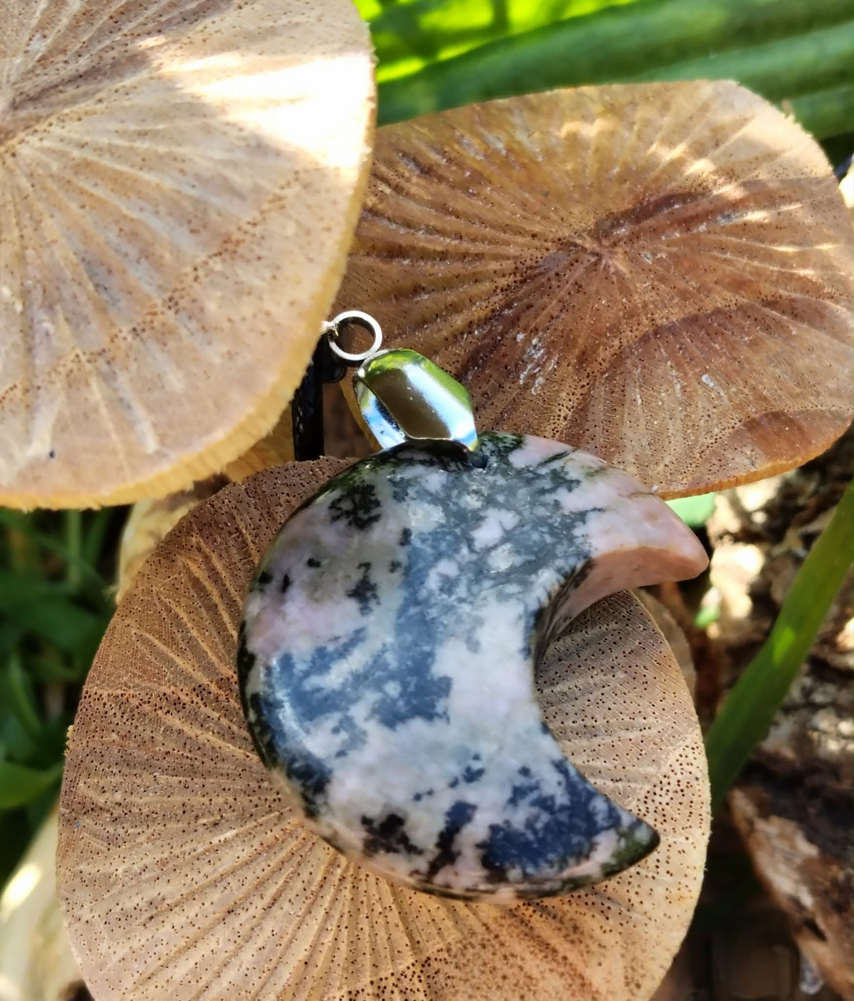 Pendentif lune en rhodonite rose marbrée de noir, posé sur champignons beige avec feuillage vert en fond, bijou en pierre naturelle associé au réconfort affectif, à la paix intérieure et à la douceur relationnelle.