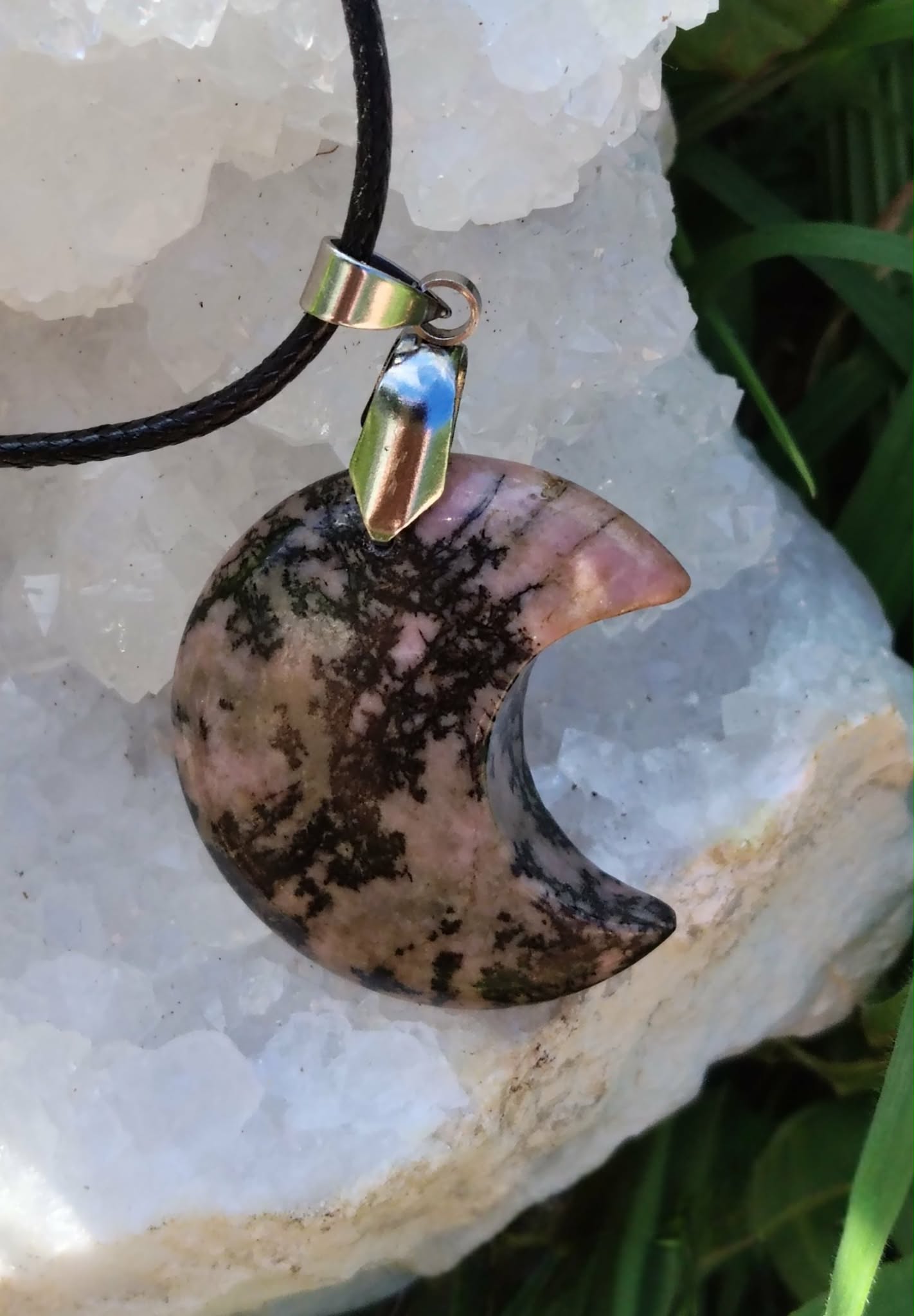 Pendentif lune en rhodonite aux nuances rose et noir, suspendu sur géode blanche avec corde et feuillage, bijou en pierre naturelle évoquant le réconfort, la tendresse et la stabilité émotionnelle.