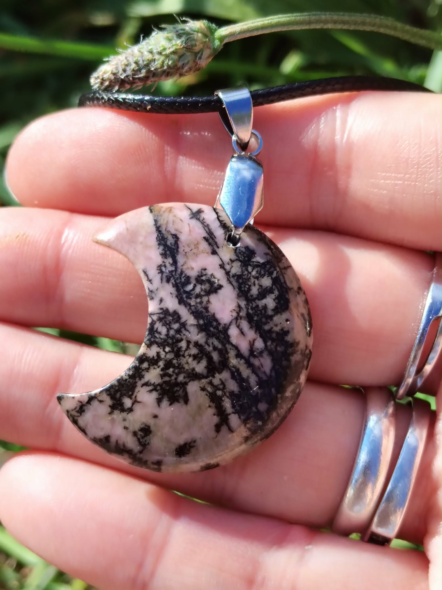 Pendentif lune en rhodonite rose marbrée de noir tenu dans une main, cordon noir et bélière argentée visibles, bijou en pierre naturelle relié à la bienveillance, au pardon et à l’équilibre du cœur.