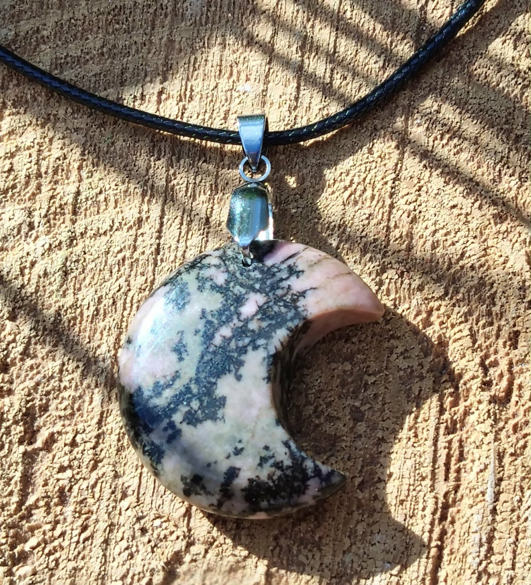 Pendentif lune en rhodonite marbrée noir et rose sur rondin de bois strié, cordon noir et bélière argentée, bijou en pierre naturelle évoquant la tendresse, la stabilité émotionnelle et l’écoute de soi.