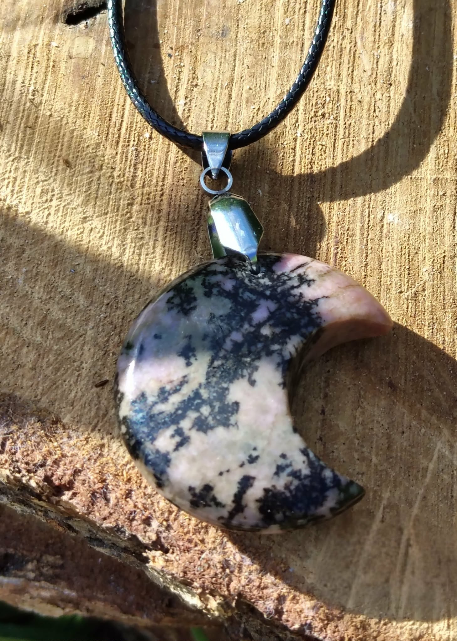 Pendentif lune en rhodonite sur cordon noir, posé sur bois brut avec ombre douce, bijou en pierre naturelle associé à la douceur du cœur, au recentrage et à l’harmonie émotionnelle.