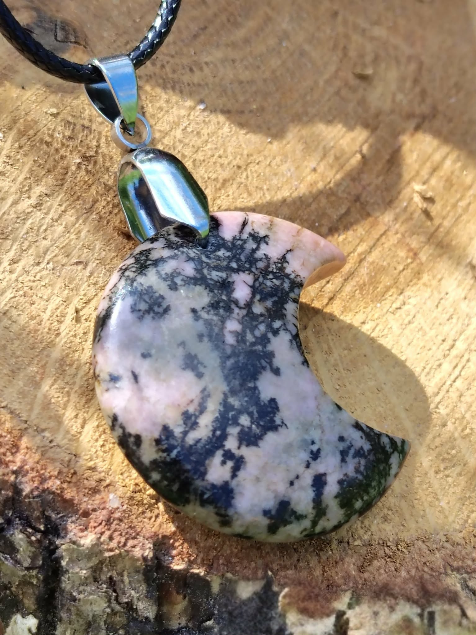 Pendentif lune en rhodonite rose veinée de noir sur cordon, posé sur une tranche de bois avec feuillage en arrière-plan, bijou en pierre naturelle lié à l’ancrage doux, au calme du cœur et au soutien émotionnel.