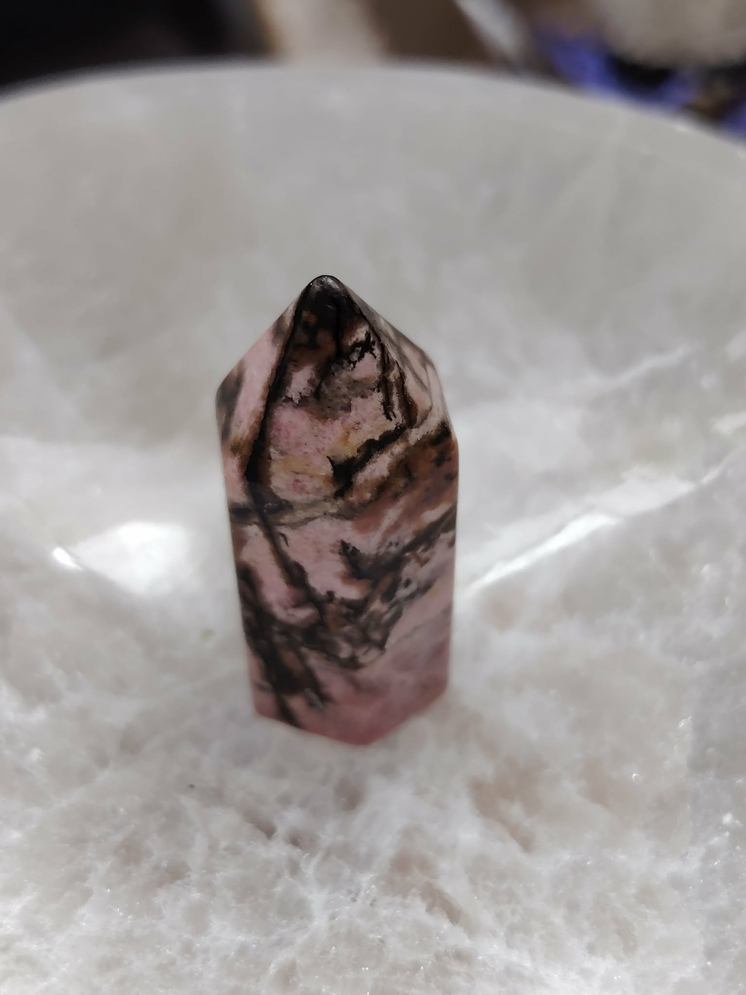 Pointe de rhodonite rose veinée de noir posée dans un bol en sélénite blanche, lumière douce, pour soutenir l’apaisement et l’équilibre émotionnel.