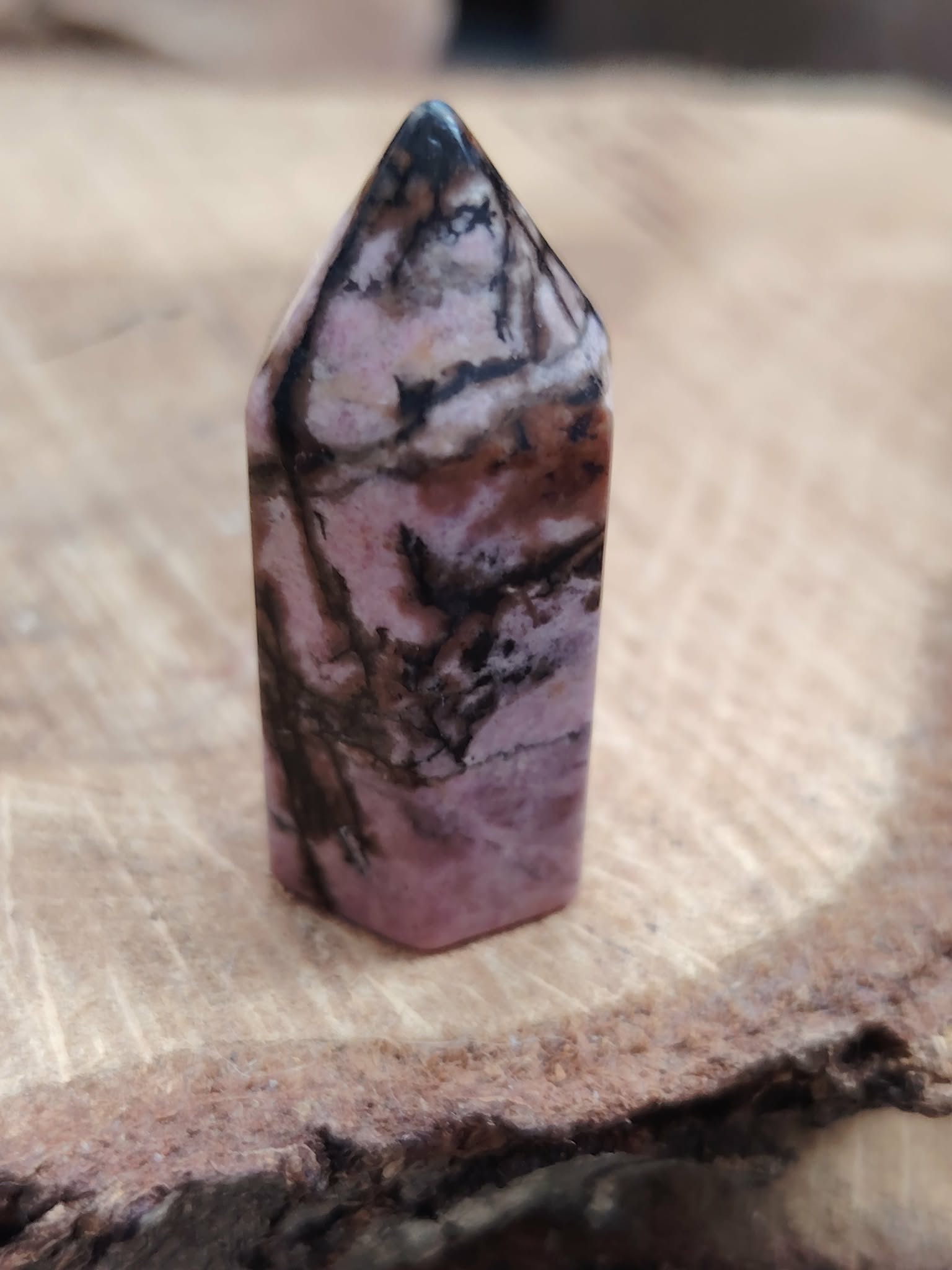 Pointe de rhodonite rose veinée de noir posée sur une tranche de bois, lumière douce et fond flou, pour apaiser le cœur et équilibrer les émotions.