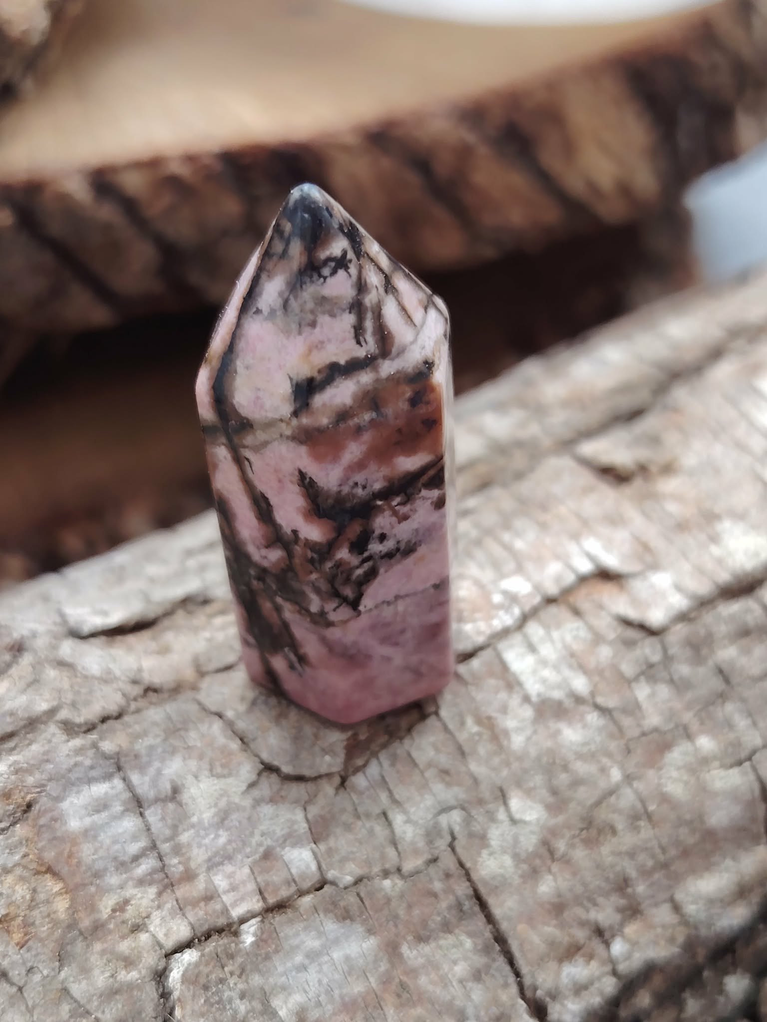 Pointe de rhodonite polie aux veines noires posée sur une écorce de bois, décor nature brun doré en lumière douce, idéale pour accompagner l’apaisement et la compassion au quotidien.