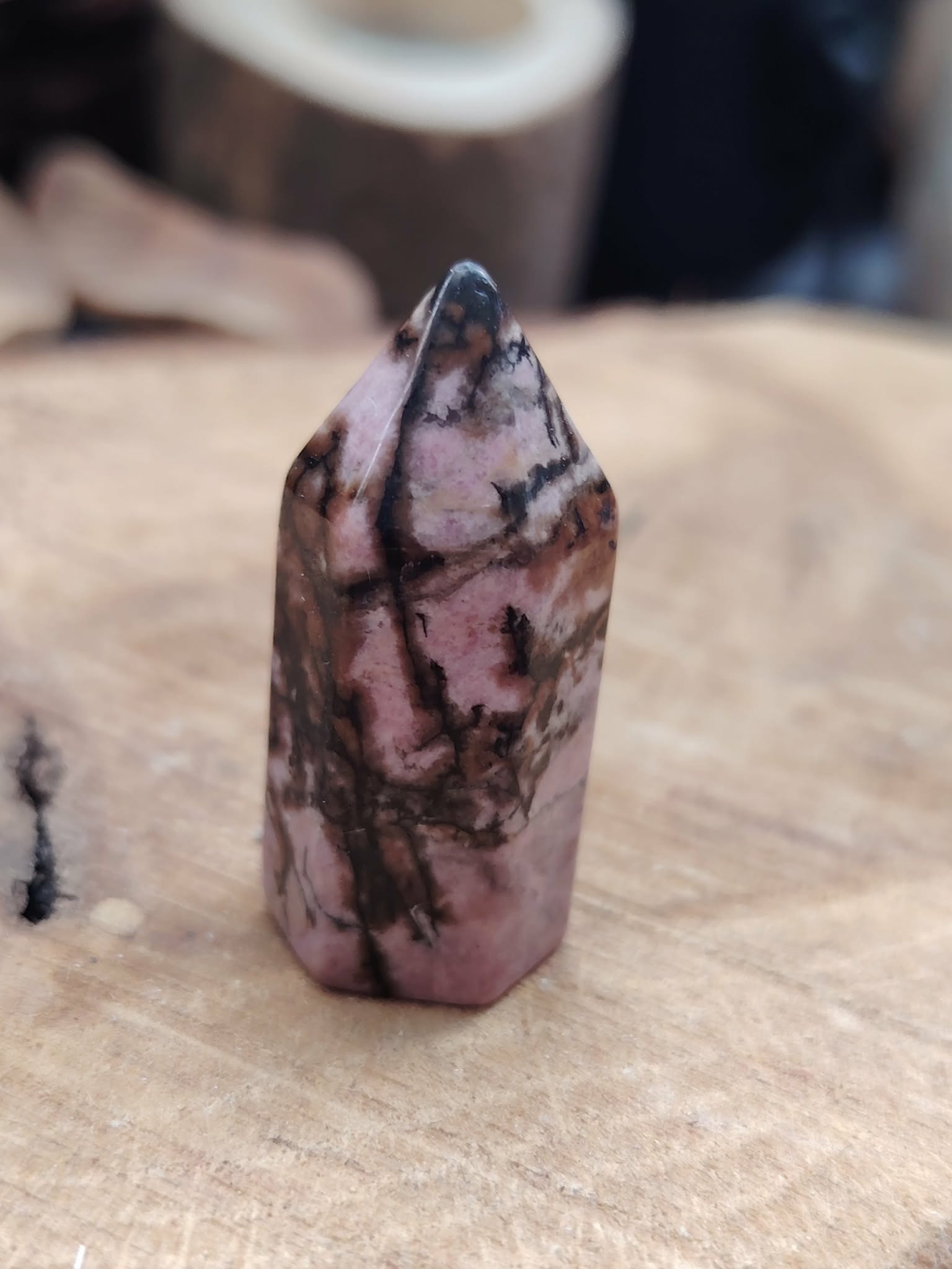 Pointe de rhodonite rose marbrée de veines noires posée debout sur une planche de bois clair, fond flou chaleureux, pour inviter à l’apaisement et à la douceur du cœur.