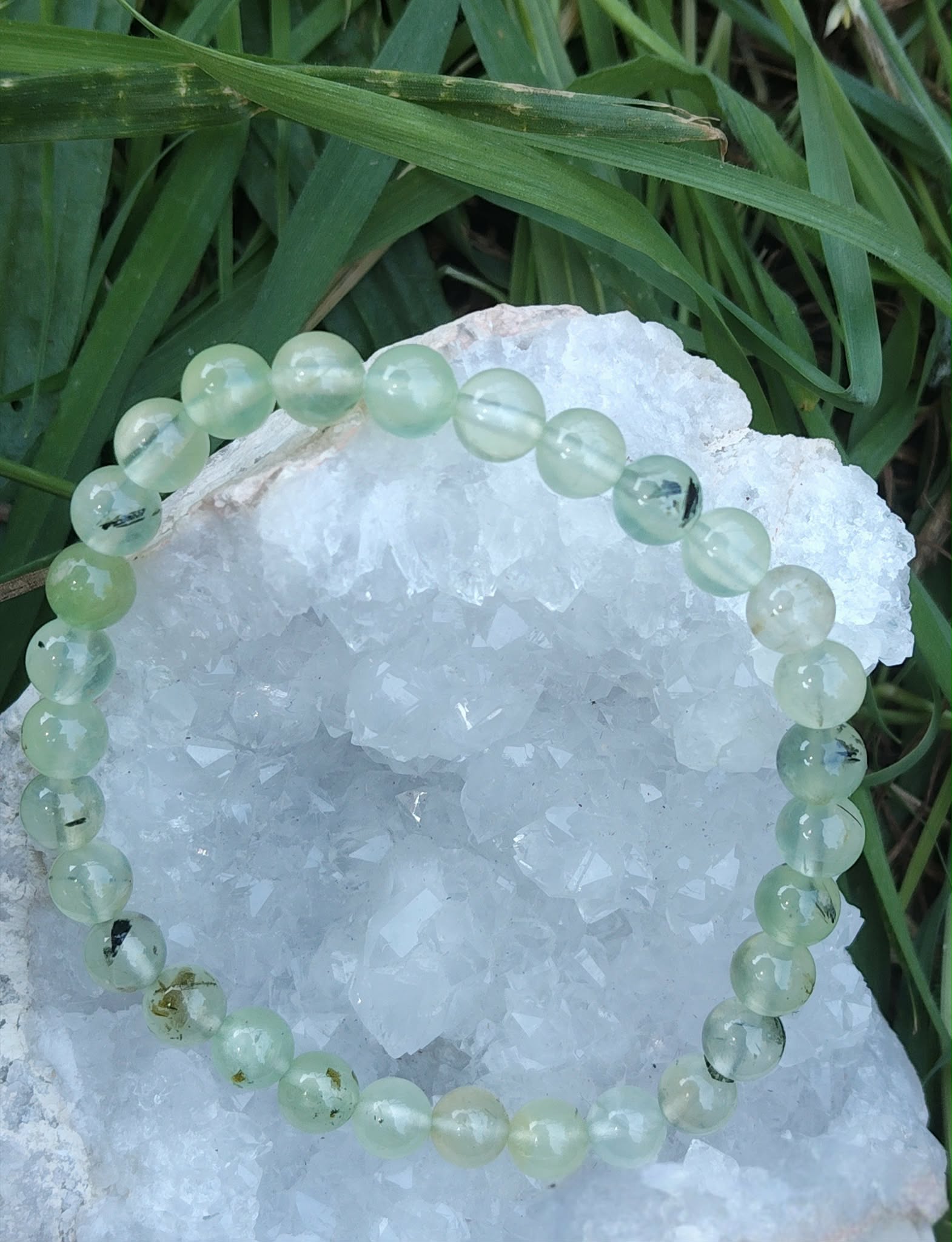 Bracelet en préhnite 6 mm vert d’eau posé sur un amas de quartz blanc, herbes vertes floues en arrière-plan, pour apaiser et clarifier l’esprit.