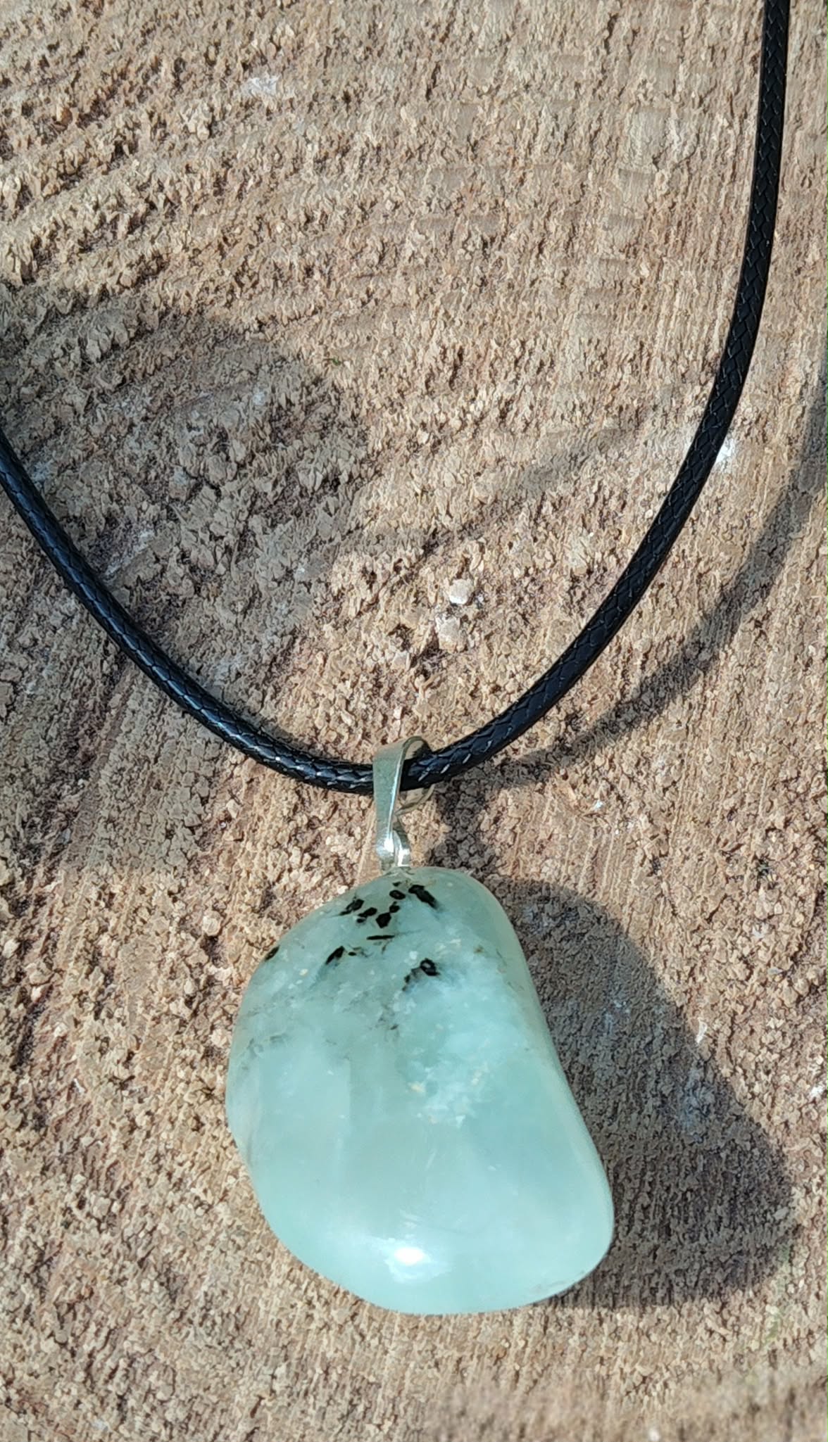 Pendentif en préhnite vert d’eau sur cordon noir, posé sur un amas de quartz blanc avec herbes en arrière-plan, pierre naturelle associée au recentrage, à la souplesse émotionnelle et à la sérénité.