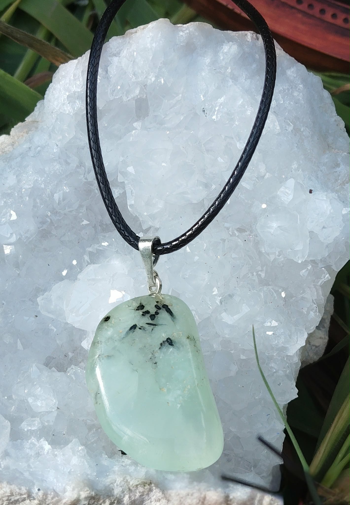 Pendentif en préhnite vert d’eau sur cordon noir, posé sur un amas de quartz scintillant parmi des brins d’herbe, bijou naturel évoquant la fraîcheur émotionnelle, le relâchement et la paix mentale.
