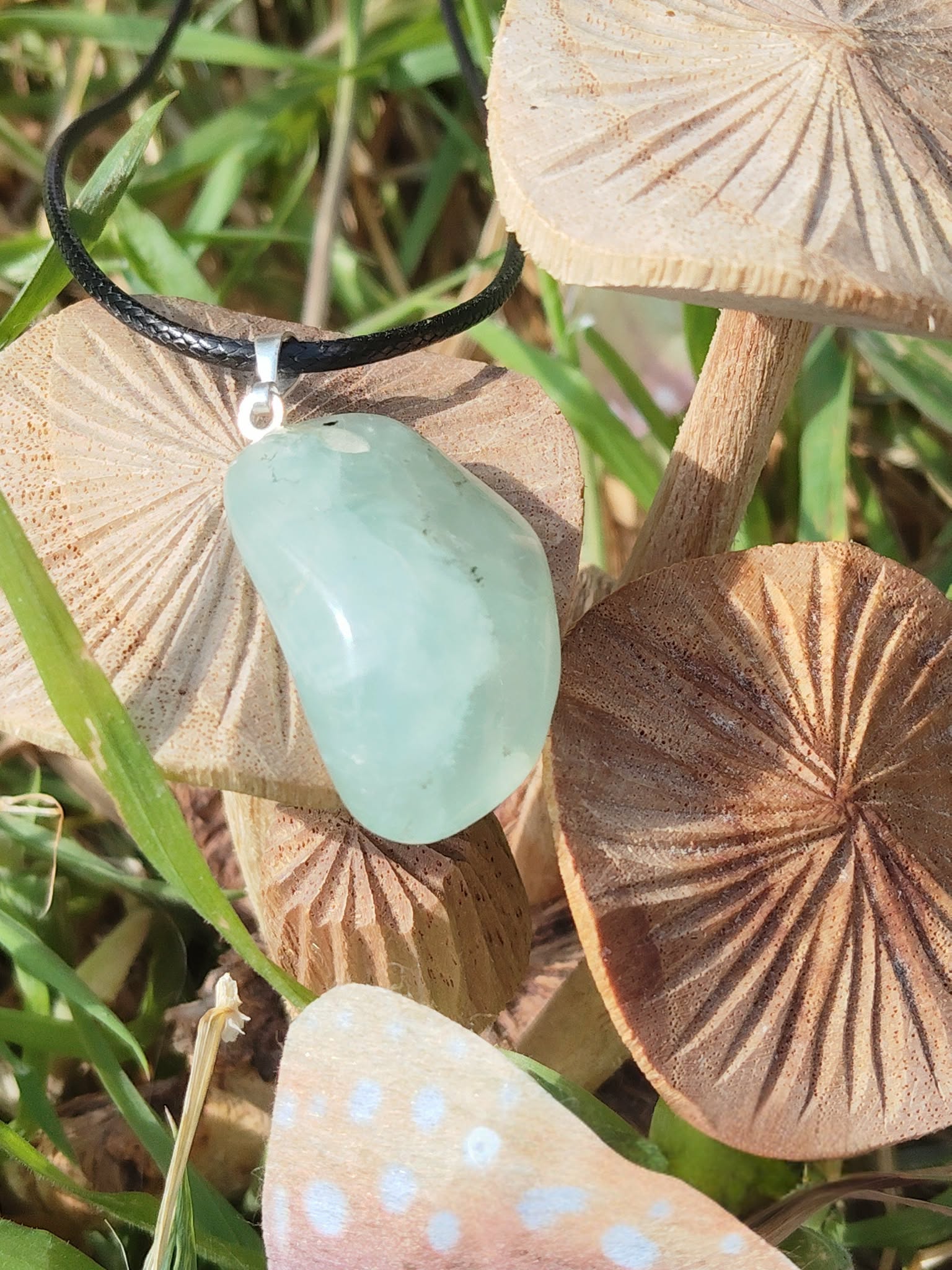 Pendentif en préhnite vert d’eau sur cordon noir, posé sur tranches de bois façon champignons au milieu d’herbes, bijou en pierre naturelle lié à la légèreté, à la respiration intérieure et à l’harmonie du cœur.