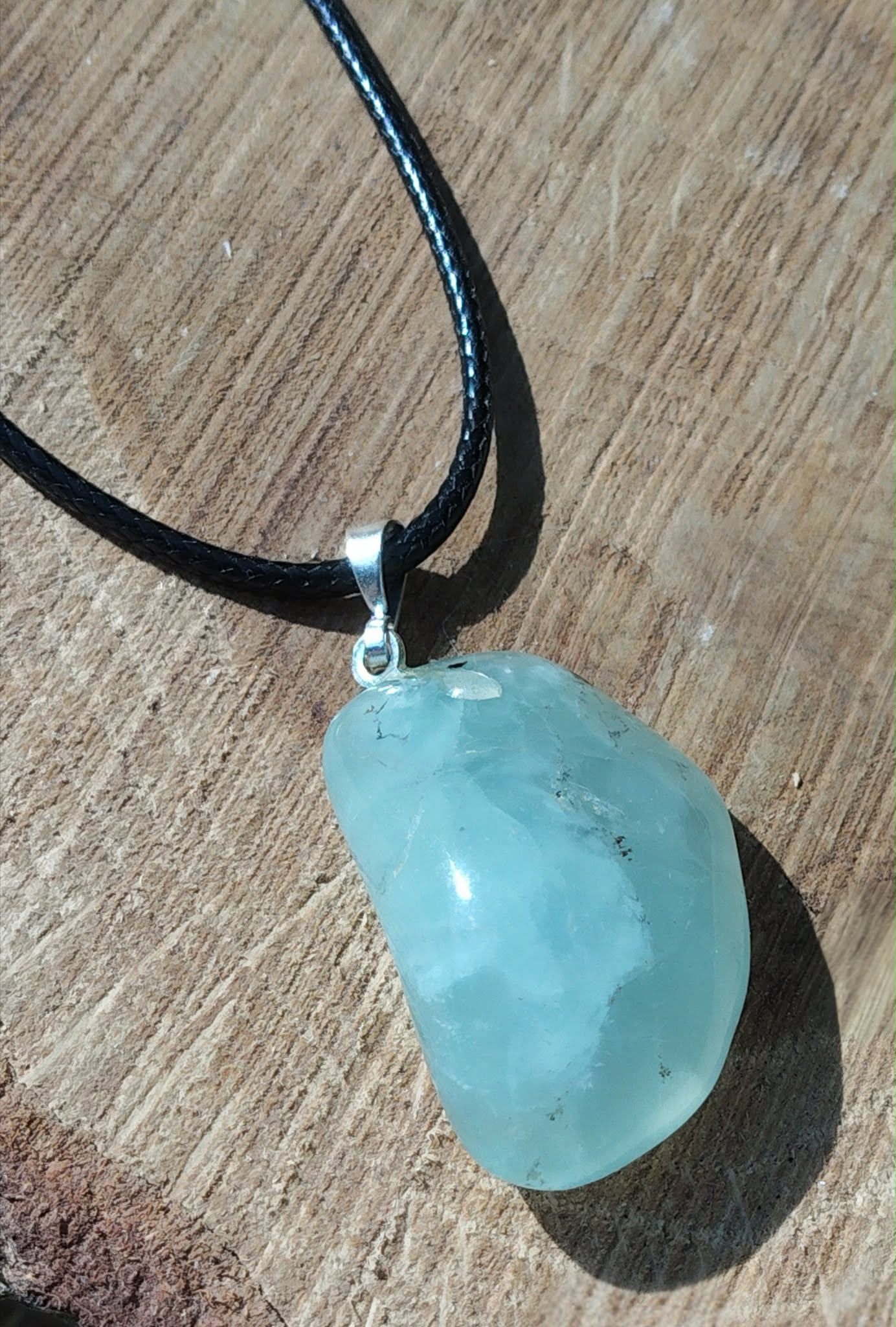 Pendentif préhnite vert d’eau sur cordon noir avec bélière argentée, photographié sur planche de bois, pierre naturelle évoquant la tranquillité, la présence à soi et l’équilibre émotionnel.