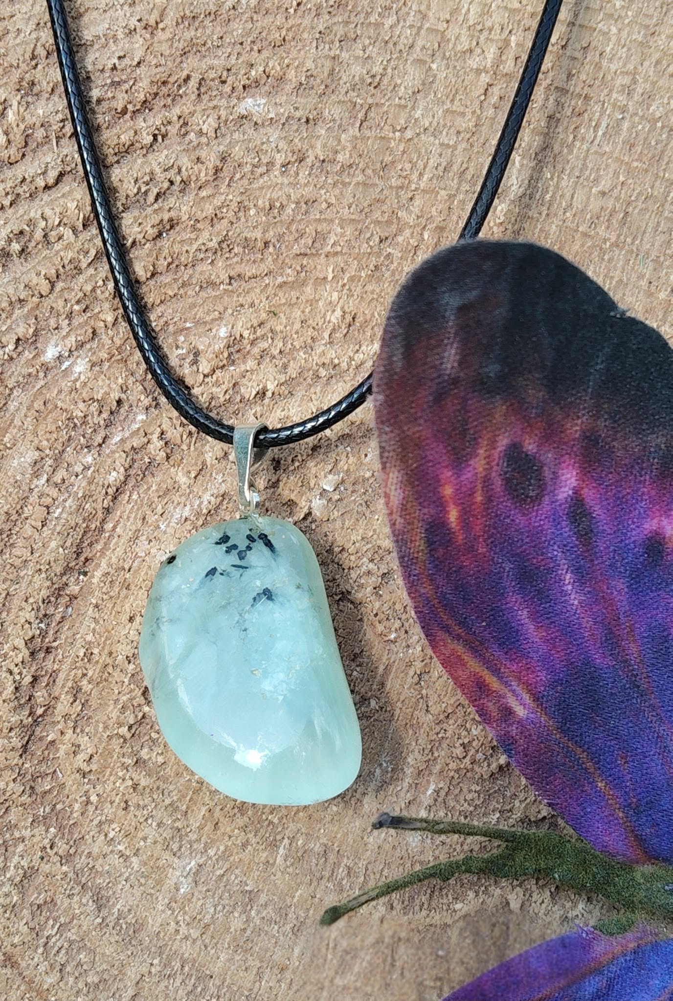 Pendentif préhnite vert d’eau sur cordon noir, posé sur tranche de bois avec papillons violets et brins d’herbe, bijou en pierre naturelle associé à la douceur, au renouveau et à l’écoute de soi.