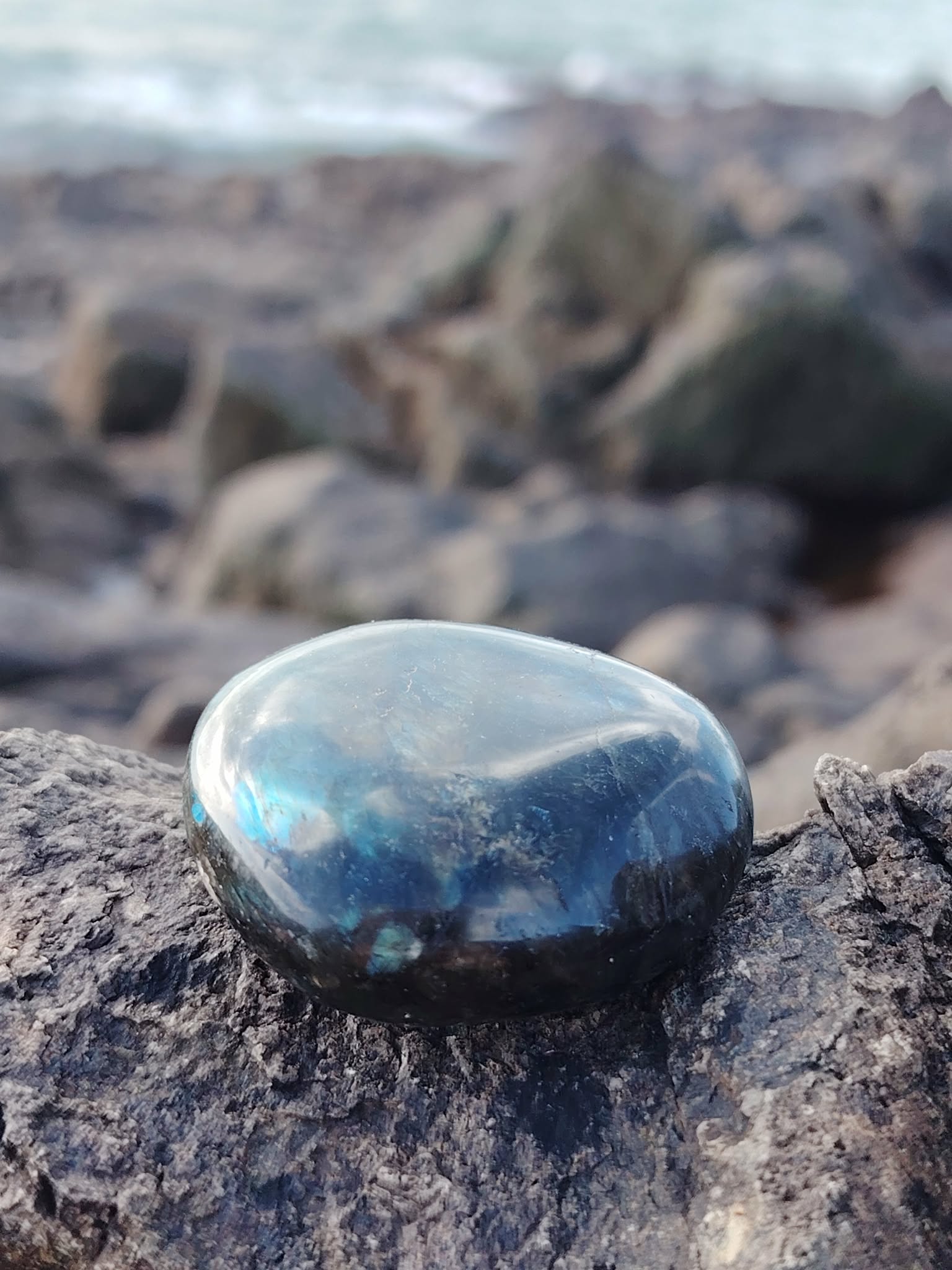 Galet de labradorite irisé bleu posé sur rocher sombre au bord de mer en Bretagne, pierre naturelle associée à l’ancrage, à la clarté d’esprit et à la protection subtile.