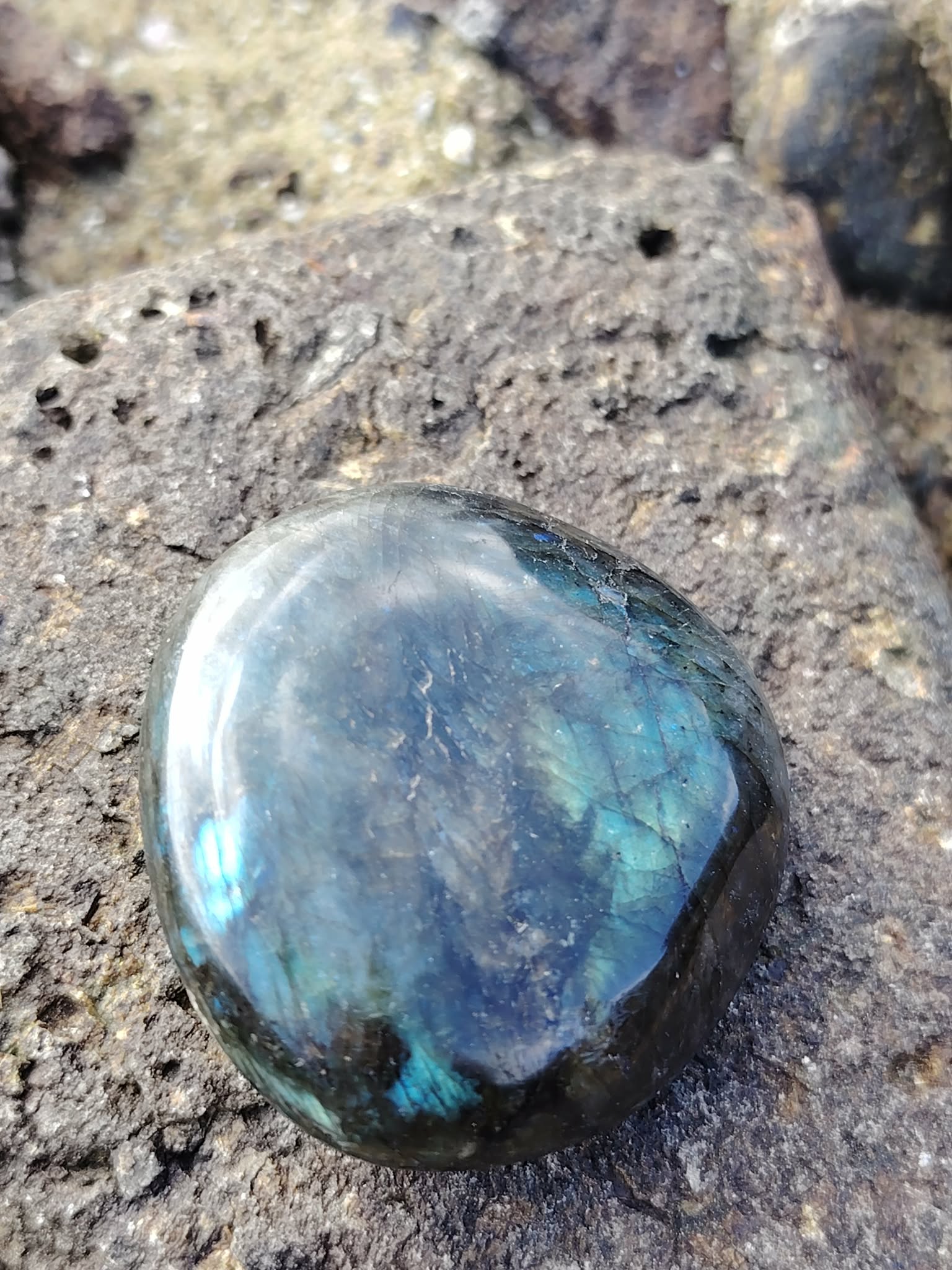 Galet de labradorite aux éclats bleu-vert posé sur roche granitique en bord de mer breton, pierre naturelle associée à la présence à soi, à la lucidité et à l’harmonie intérieure.
