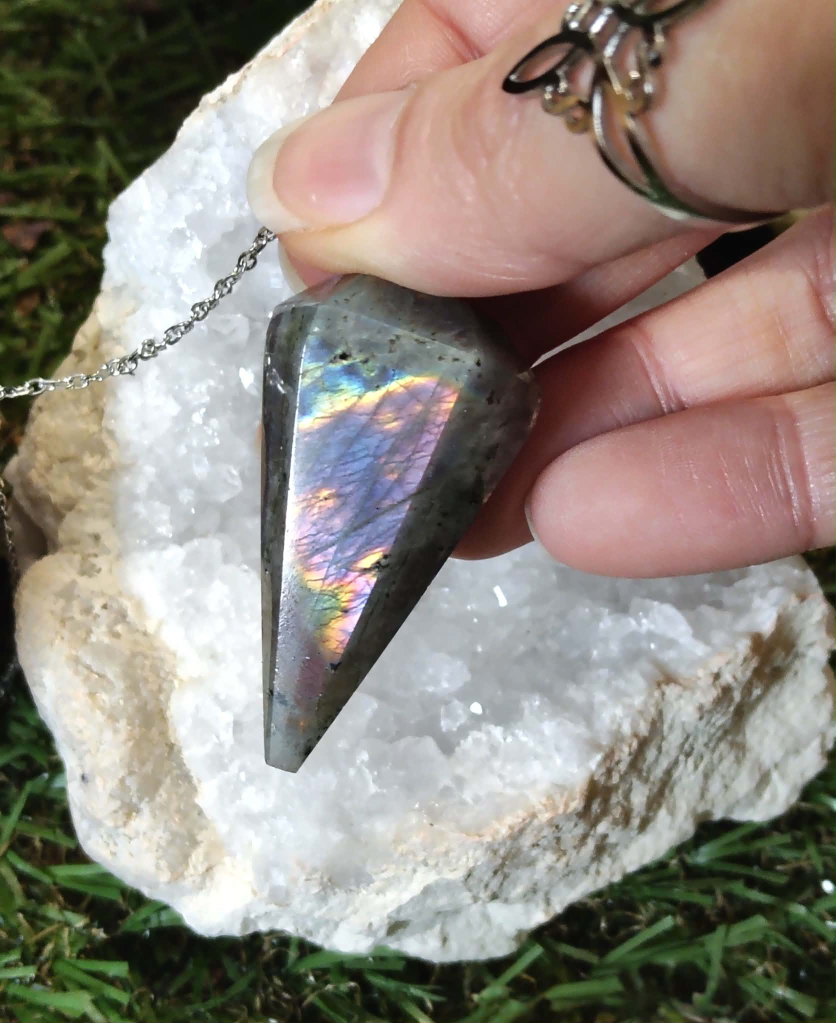 Pendule en labradorite arc-en-ciel tenu entre les doigts, posé sur un amas de quartz blanc et mousse verte, pour se recentrer, se protéger et écouter son intuition.