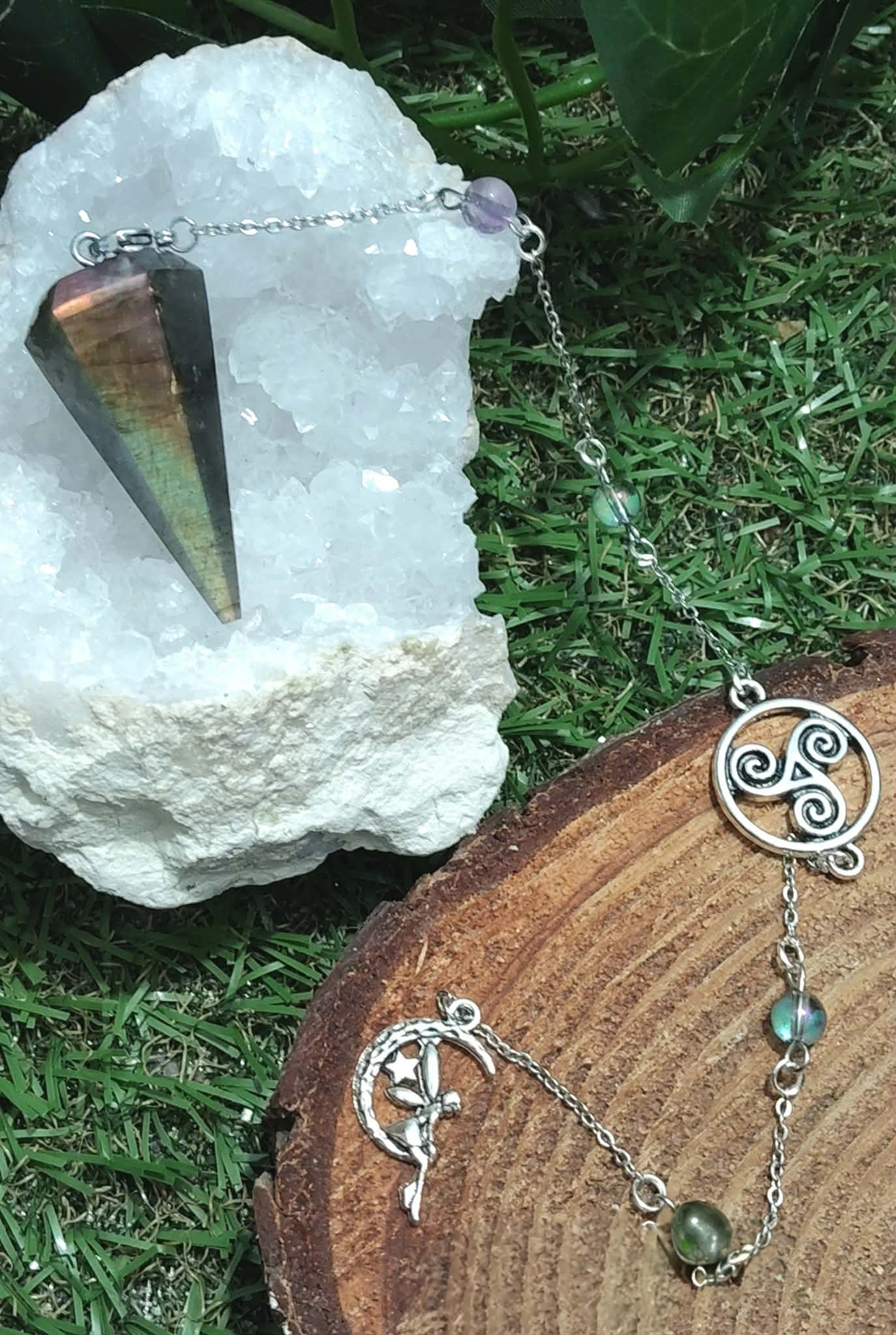 Pendule conique en labradorite arc-en-ciel posé sur un amas de quartz, sur mousse verte et rondin de bois avec breloques, pour affiner l’intuition et se recentrer.