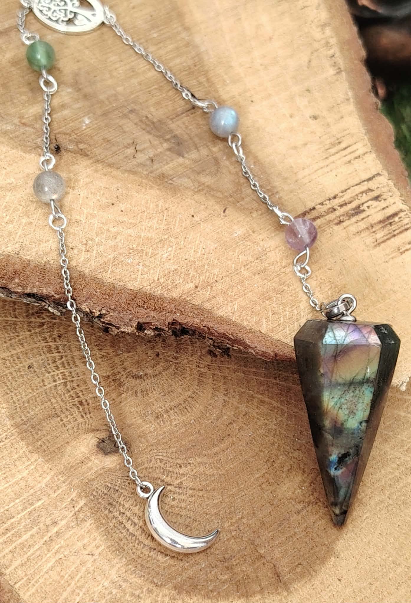 Pendule en labradorite aux reflets irisés, chaînette argentée avec perles et lune, posé sur une tranche de bois, pour protéger l’énergie et affiner l’intuition