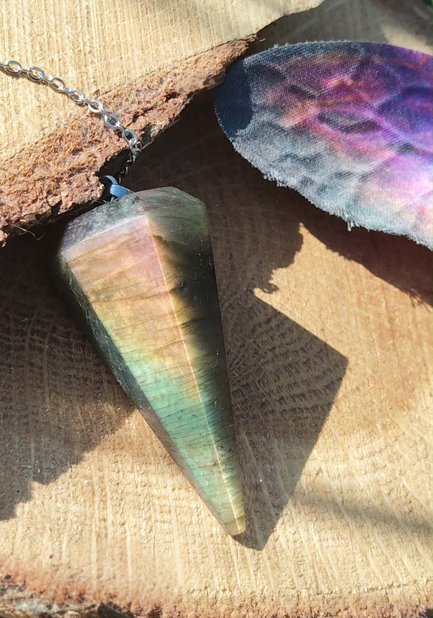 Pendule conique en labradorite aux reflets arc-en-ciel posé sur une tranche de bois, près d’un papillon irisé et de mousse verte, pour se recentrer et affiner l’intuition.