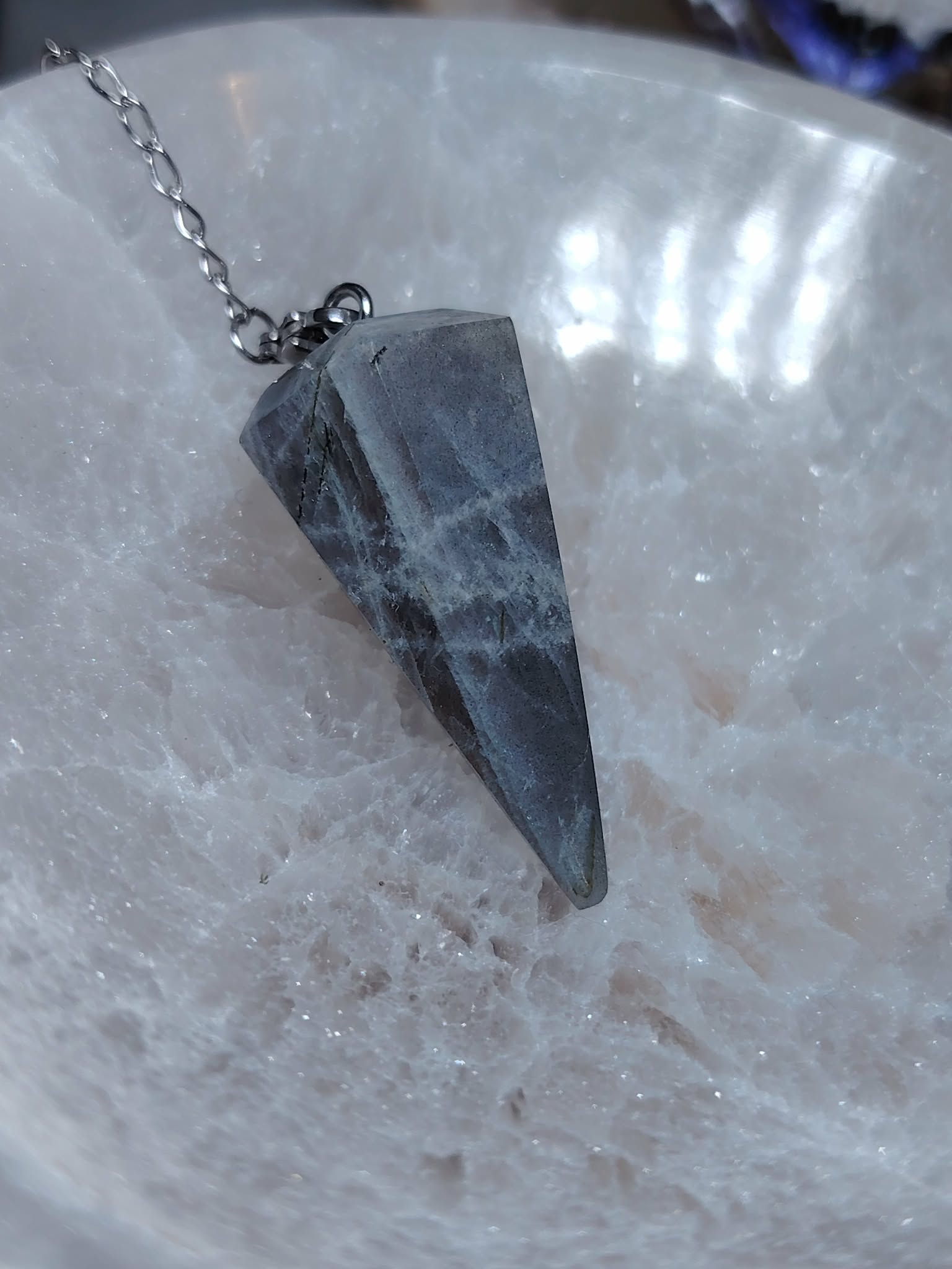 Pendule en labradorite sur chaîne argentée, posé sur une plaque de sélénite blanche, reflets doux, pour protection, intuition et clarté.