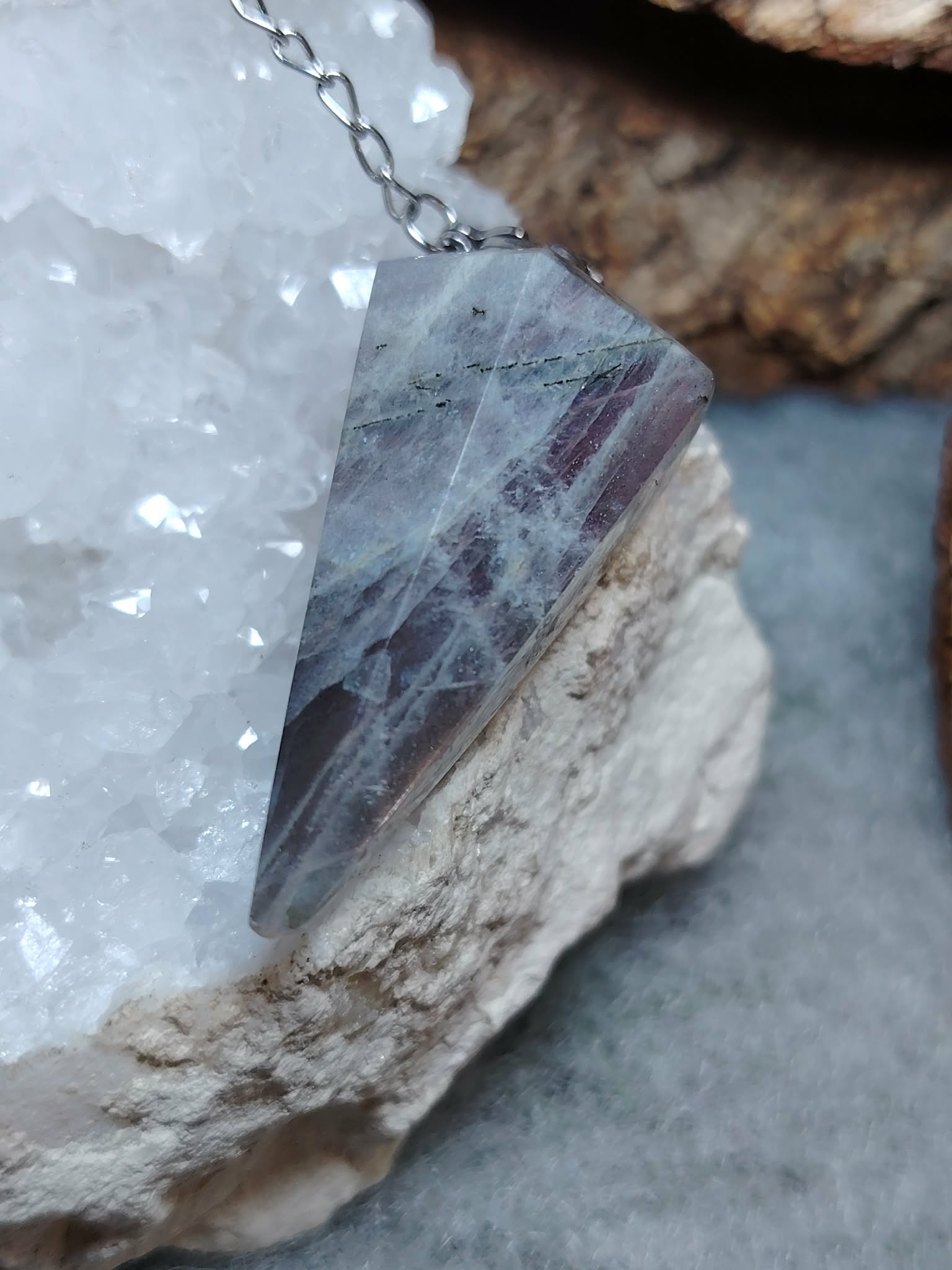Pendule en labradorite posé sur un amas de quartz, sur fond gris avec une écorce, reflets bleus pour protection et clarté intérieure.