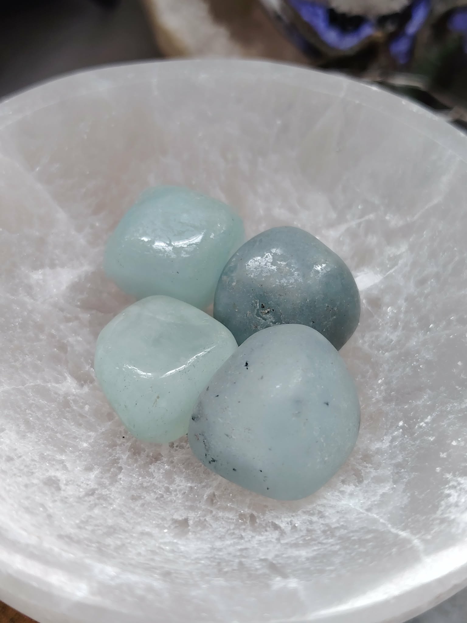 Pierres roulées d’aigue-marine bleu doux posées dans un bol en sélénite blanche, décor naturel, pour une sensation de calme, clarté et expression fluide.