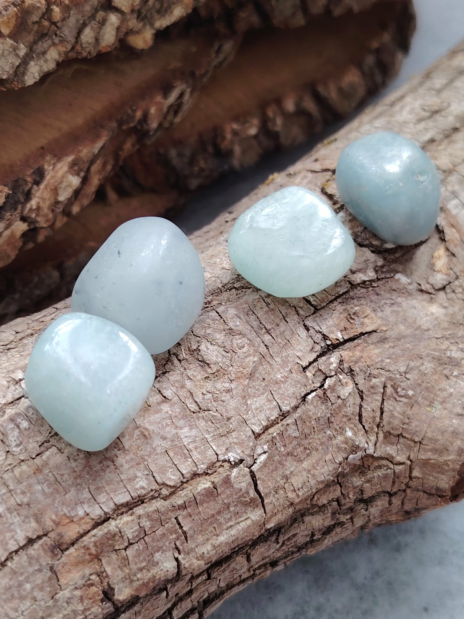 Quatre pierres roulées d’aigue-marine bleu pastel posées sur écorce et bois craquelé, ambiance nature, pour apaiser le mental et faciliter une communication douce.