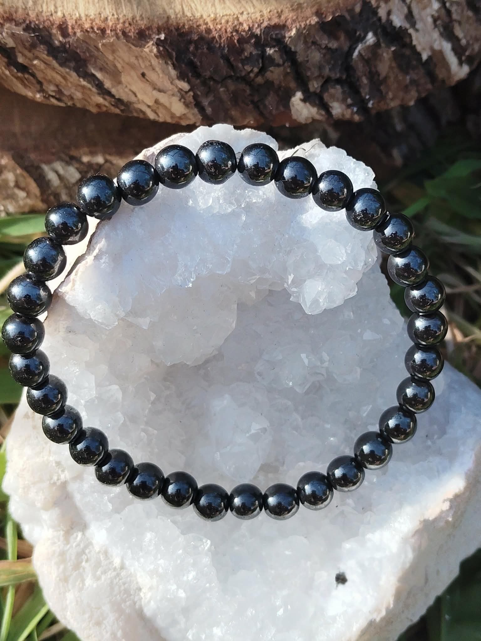 Bracelet hématite 6 mm posé sur bloc de quartz blanc avec écorce et herbes, perles gris acier pour maîtrise de soi, stabilité et contrôle calme