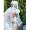 Spell jar en fiole de verre avec cristal de roche, fil rouge et cire blanche, posé sur bois avec coquillage et herbe en fond, ambiance protection et purification.