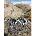 Boucles d’oreilles Magic Gaïa en larimar et aqua aura posées sur un rocher granitique en Bretagne, face à la mer floue, avec perles bleu ciel et blanches, triquetra argentée, évoquant douceur, intuition et apaisement.