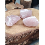 Trois quartz rose bruts posés sur tranche de bois avec galets beiges en arrière-plan, pierres naturelles associées à la douceur relationnelle, à la sérénité et à l’harmonie émotionnelle.