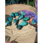 Galets de chrysocolle bleu-vert sur tranche de bois avec plume violette et feuillage, pierres roulées associées à l’intuition douce, à la sensibilité apaisée et à la parole fluide.