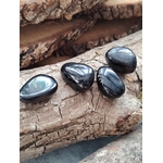 Quatre pierres roulées d’hyperstène noir aux reflets métalliques posées sur du bois craquelé, lumière douce, pour inviter à l’ancrage, la protection et la clarté mentale