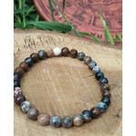 Bracelet Piétersite 6 mm création Magic Gaïa, perles brun-bleu nuancées en cercle sur rondin de bois gravé, herbes vertes et écorce en fond; apporte ancrage, clarté et courage.