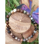 Bracelet en piétersite 6 mm aux perles brun, bleu et or, posé sur rondin de bois gravé, herbes vertes et papillon violet en décor; apporte ancrage, clarté et confiance.