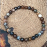 Bracelet piétersite 6 mm aux perles bleu pétrole, brun et doré, posé sur tissu sable près d’écorce et d’herbes vertes; invite à la clarté, au courage et à l’ancrage.