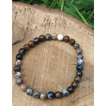 Bracelet Piétersite 6 mm en perles brun-bleu, posé en cercle sur rondin de bois gravé, brin d’herbe en arrière-plan; aide à l’ancrage, lucidité et confiance