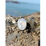 Bague réglable en argent 925 sertie d’une péristérite ovale (pierre de lune arc-en-ciel) posée sur rochers, mer de Bretagne en arrière-plan, reflets bleutés nacrés, intention apaisement et intuition.