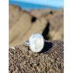 Bague réglable en argent 925 sertie d’une péristérite ovale (pierre de lune arc-en-ciel) posée sur roche granitée, mer de Bretagne floutée en arrière-plan, reflets laiteux, intention apaisement et intuition.
