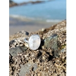 Bague réglable en argent 925 sertie d’une péristérite ovale (pierre de lune arc-en-ciel) posée sur rochers, plage et mer de Bretagne floutées en arrière-plan, reflets nacrés, intention apaisement et intuition