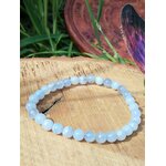 Bracelet en calcédoine bleue 6 mm sur rondin de bois clair au soleil, herbe verte, disque gravé et plume irisée, sérénité intérieure et clarté dans l’échange.