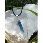 Pendentif cyanite bleue brute sur cordon noir, bélière argentée, posé sur amas de quartz blanc et herbes; intention: apaisement, expression.