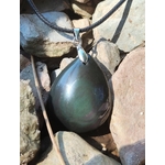 Pendentif obsidienne œil céleste en goutte sur cordon noir, posé entre galets clairs au soleil avec reflets verts et pourpres, bijou en pierre naturelle évoquant la force intérieure, l’enracinement et la vigilance.
